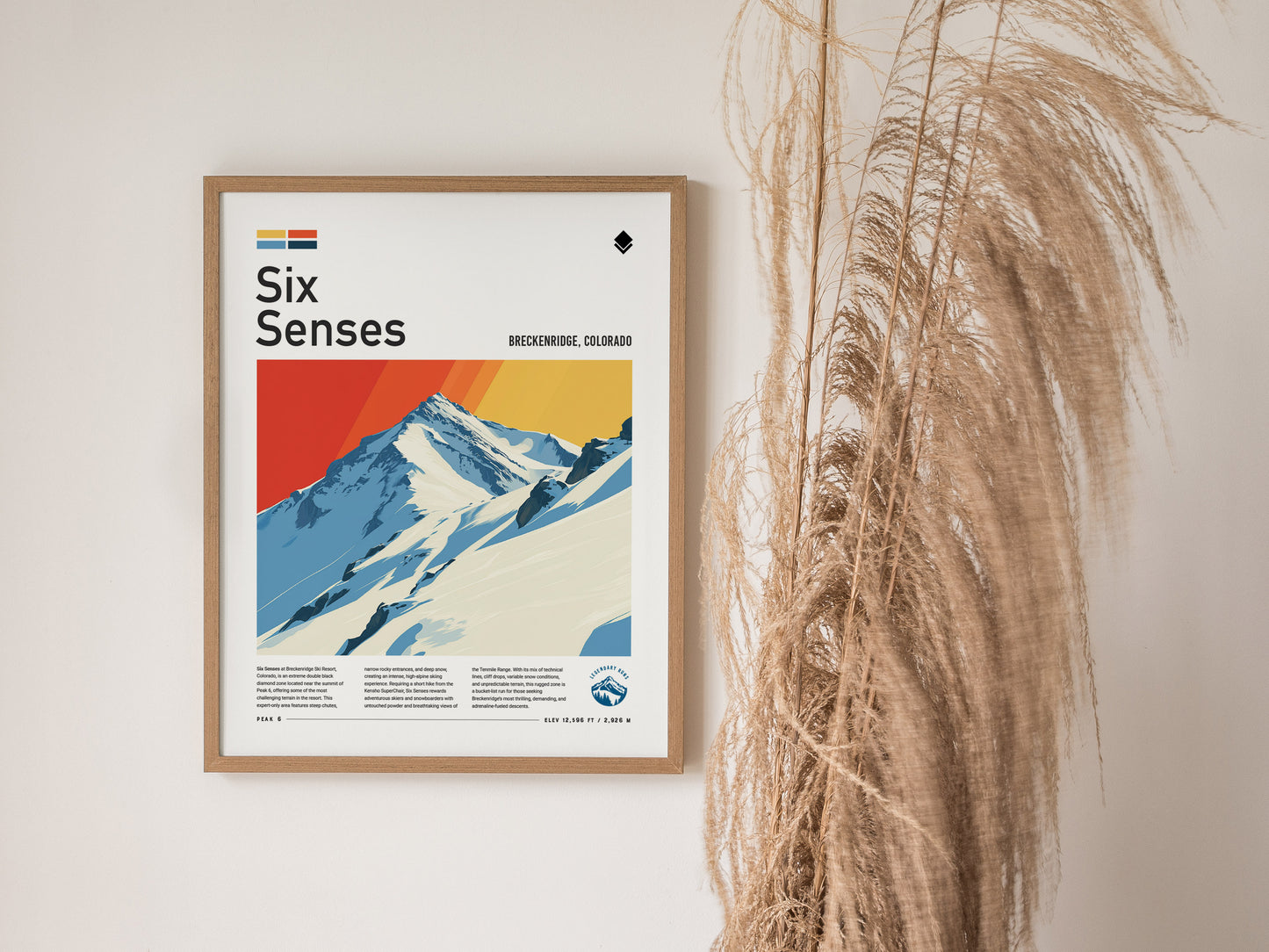 Framed Six Senses poster depicting a stylized snowy mountain peak with vibrant red and yellow sky. The background features dried ornamental grass against a light-colored wall.