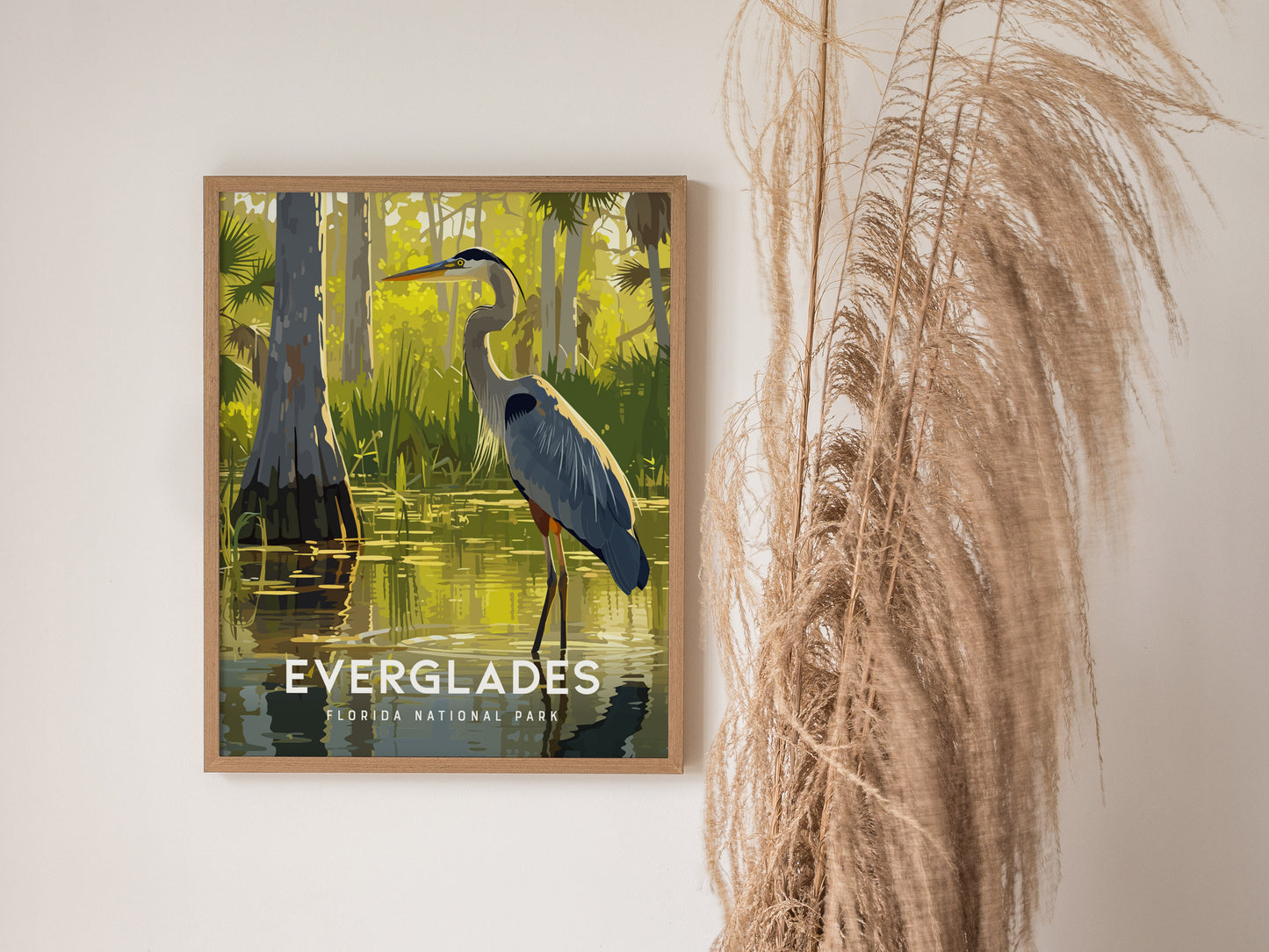 A framed poster of a Great Blue Heron in a serene Everglades scene hangs on a wall, next to tall, wispy dried grass.