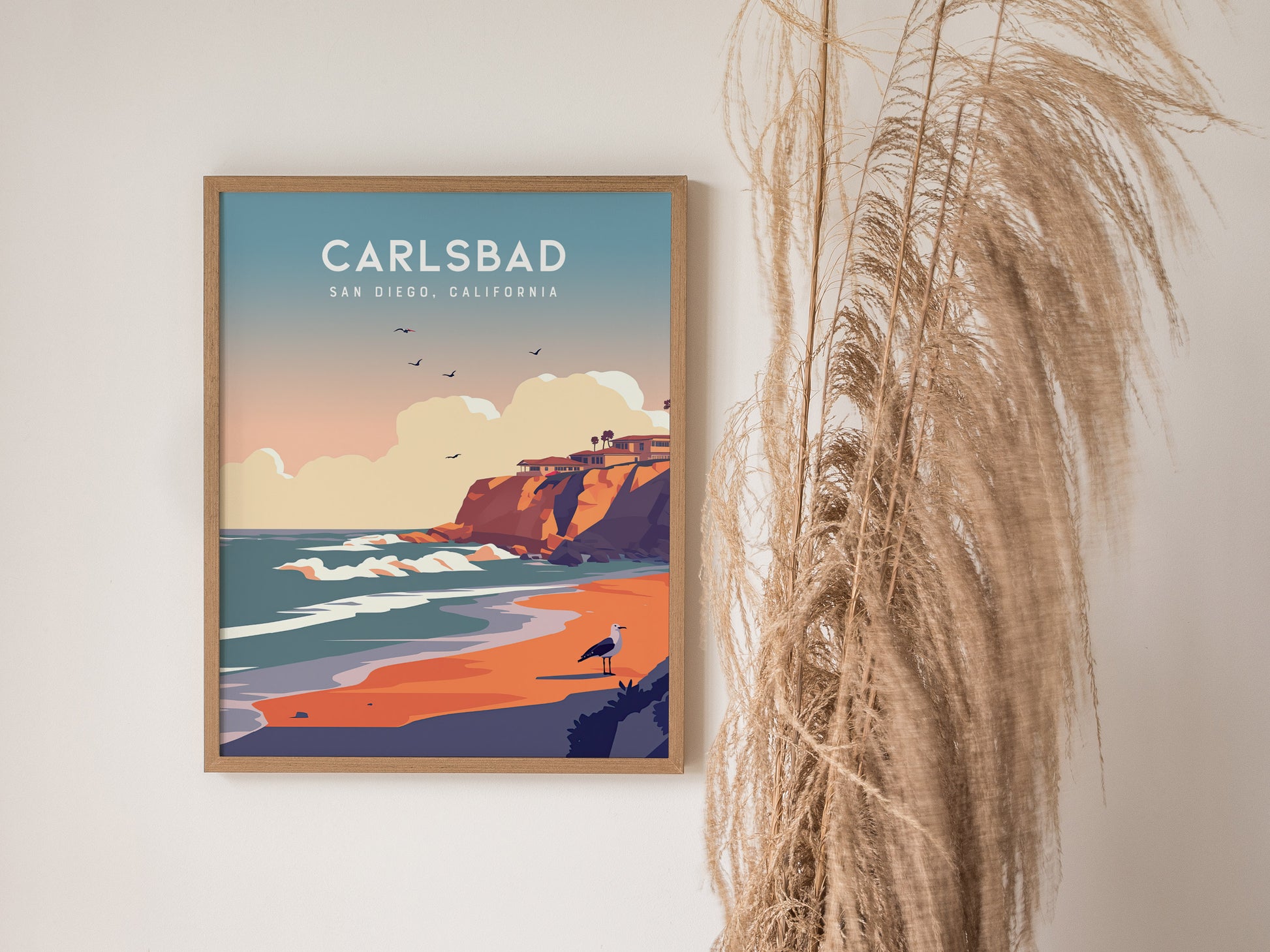 Framed poster of Carlsbad, San Diego, depicting cliffs, ocean waves, and a sandy beach with a bird, set against a pastel sky. Decorative dried grasses are beside the artwork.