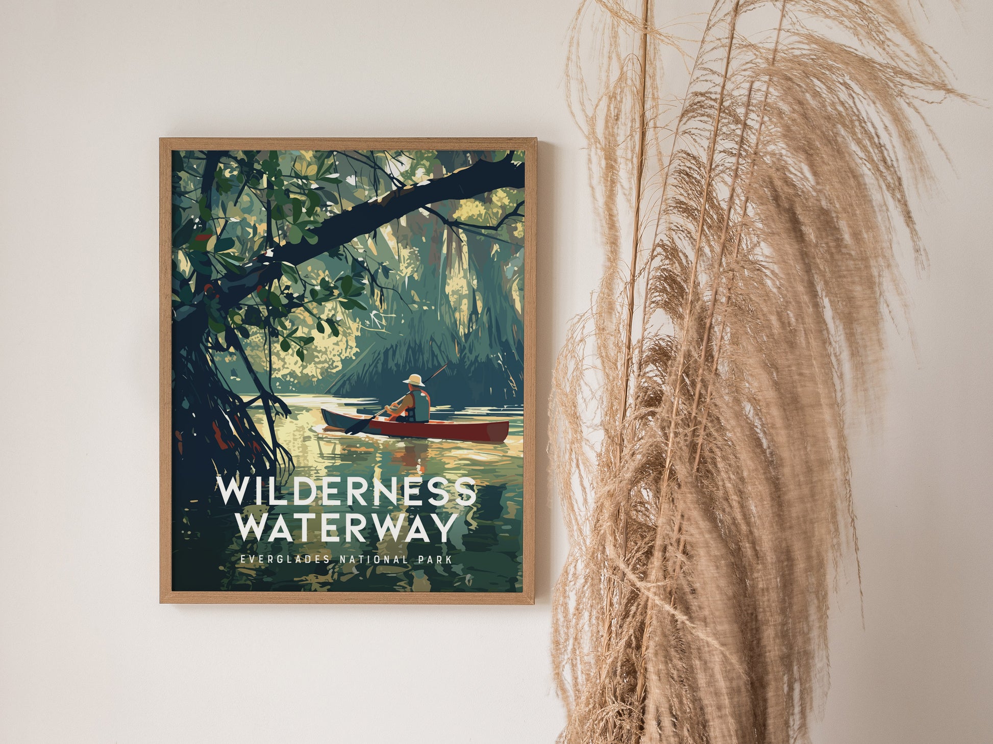 A framed Wilderness Waterway poster depicts a canoer navigating through lush mangrove forests, with decorative dried grasses placed beside it against a white wall.