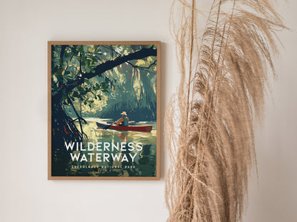 A framed Wilderness Waterway poster depicts a canoer navigating through lush mangrove forests, with decorative dried grasses placed beside it against a white wall.
