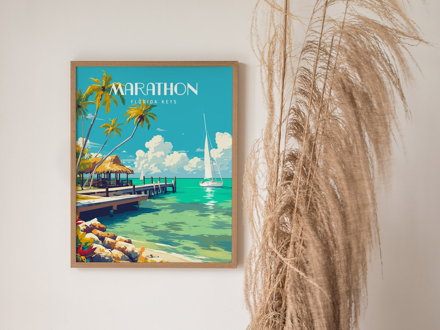 A framed poster of Marathon, Florida Keys showcases a tropical scene with palm trees, a dock, and a sailboat on turquoise water, beside dried pampas grass.