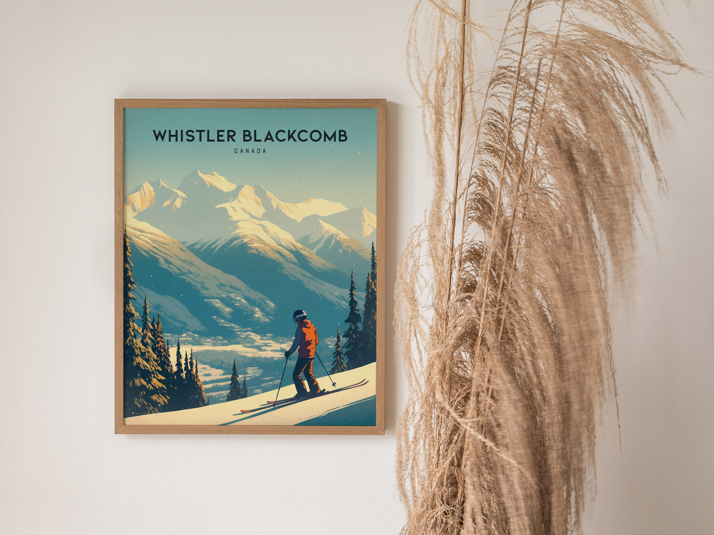 A framed poster of Whistler Blackcomb features a skier on snowy slopes with towering mountains in the background, displayed beside tall, wispy pampas grass.