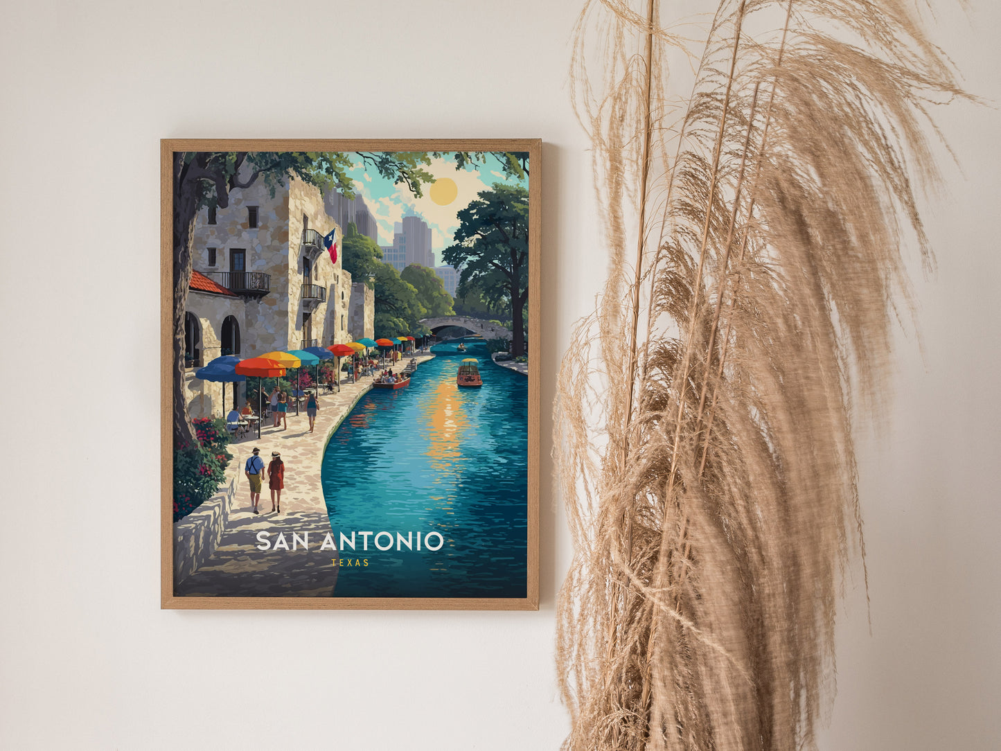 A framed poster of San Antonios riverwalk features colorful umbrellas and a tranquil river scene, with two people walking along the path. Tall dried pampas grass decorates the side.