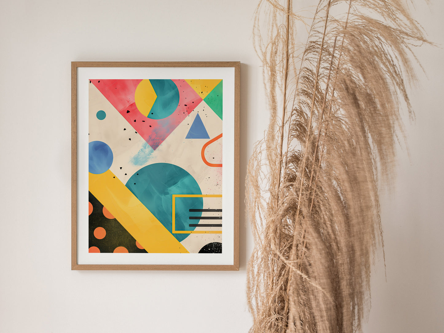 A framed abstract art piece featuring vibrant geometric shapes in pink, blue, yellow, and green hues. Its displayed on a wall beside tall, dried pampas grass.