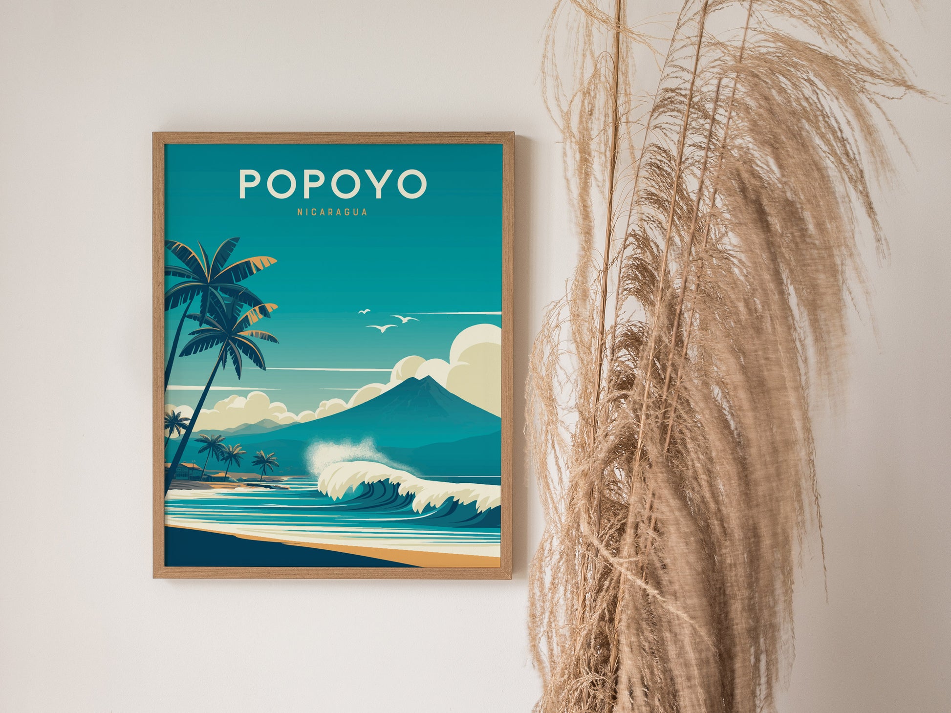 A framed Popoyo Nicaragua surf poster features palm trees, waves, and a distant volcano under a blue sky. Tall dried grasses are placed beside the artwork.