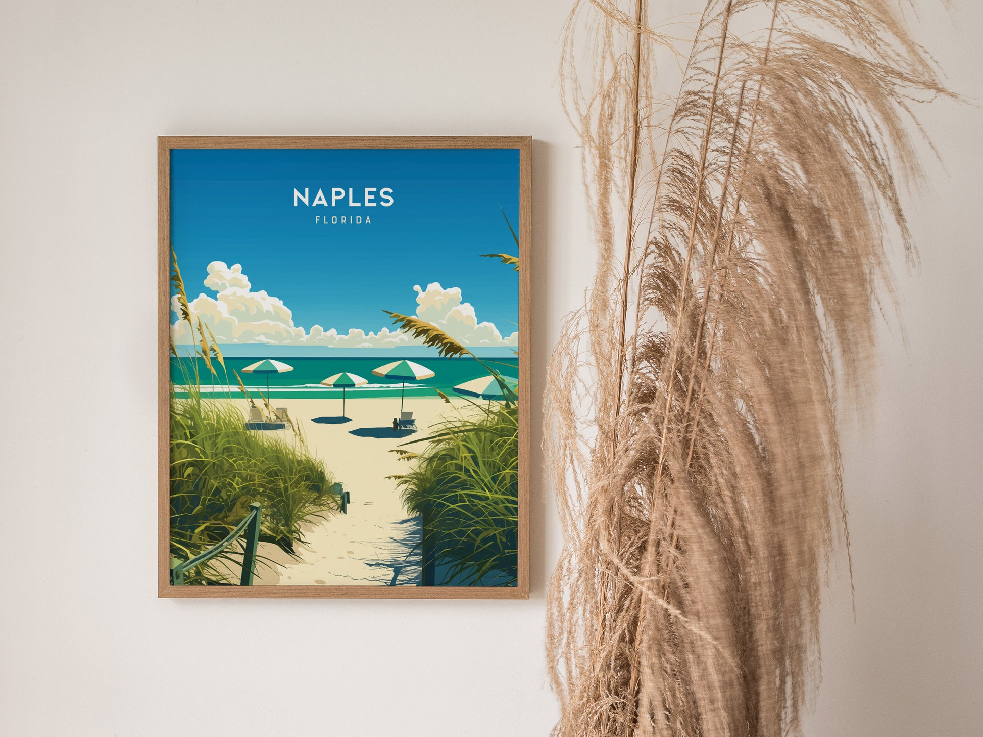 A framed poster of Naples, Florida beach scene with umbrellas and lush greenery is displayed on a wall next to tall, dried pampas grass.