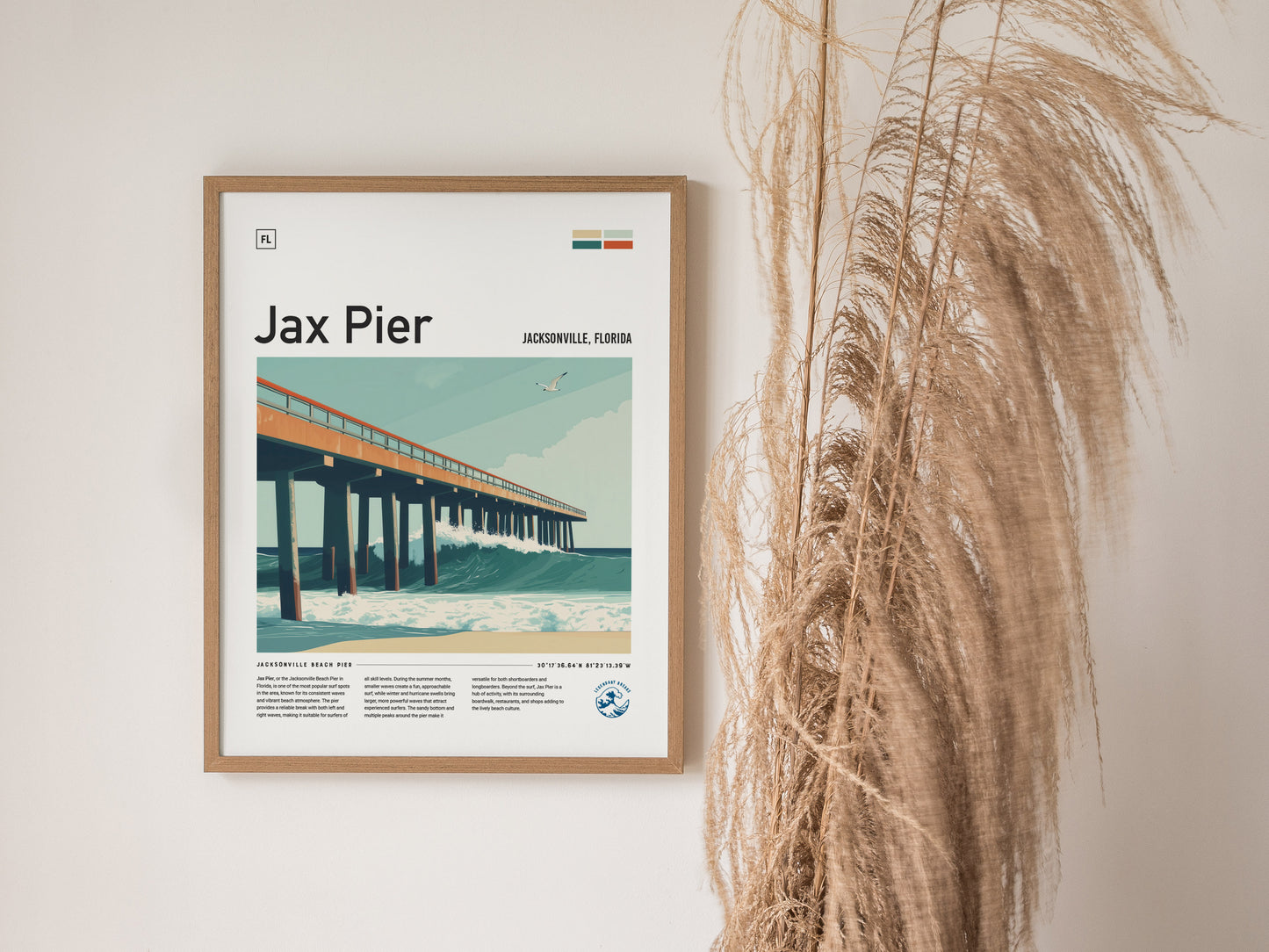 Framed artwork of Jax Pier, Jacksonville, Florida, featuring a stylized illustration of a pier over ocean waves. Tall, dried grasses are positioned beside the artwork against a light wall.