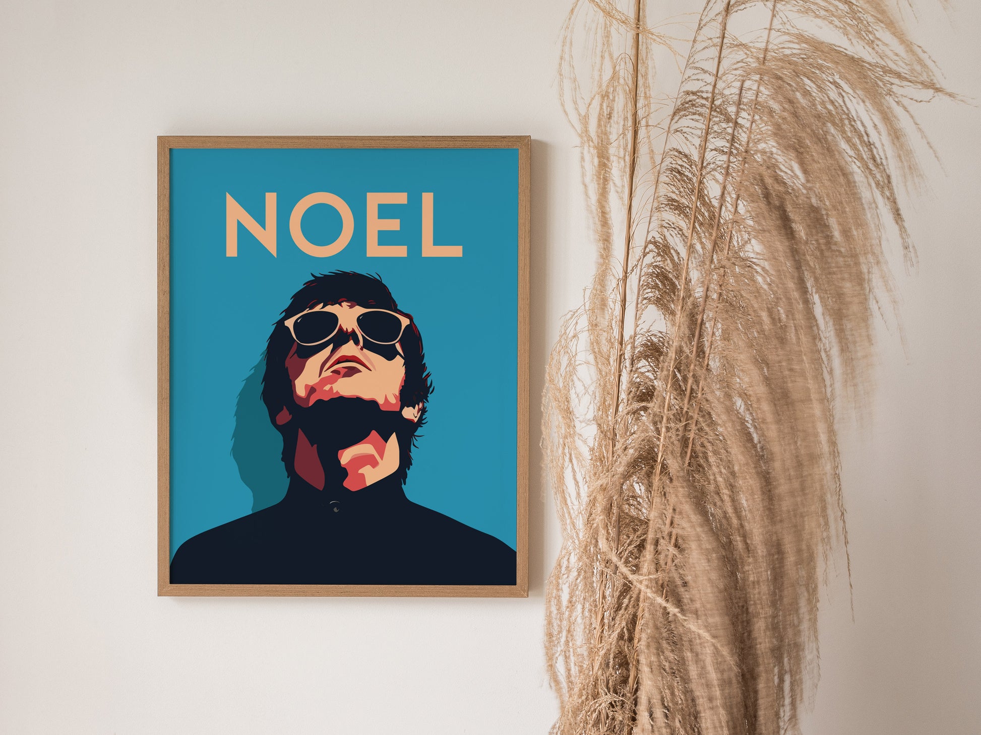 A framed poster featuring an illustrated figure with sunglasses and the word NOEL in bold text against a blue background, next to tall dried grasses.