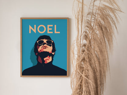 A framed poster featuring an illustrated figure with sunglasses and the word NOEL in bold text against a blue background, next to tall dried grasses.