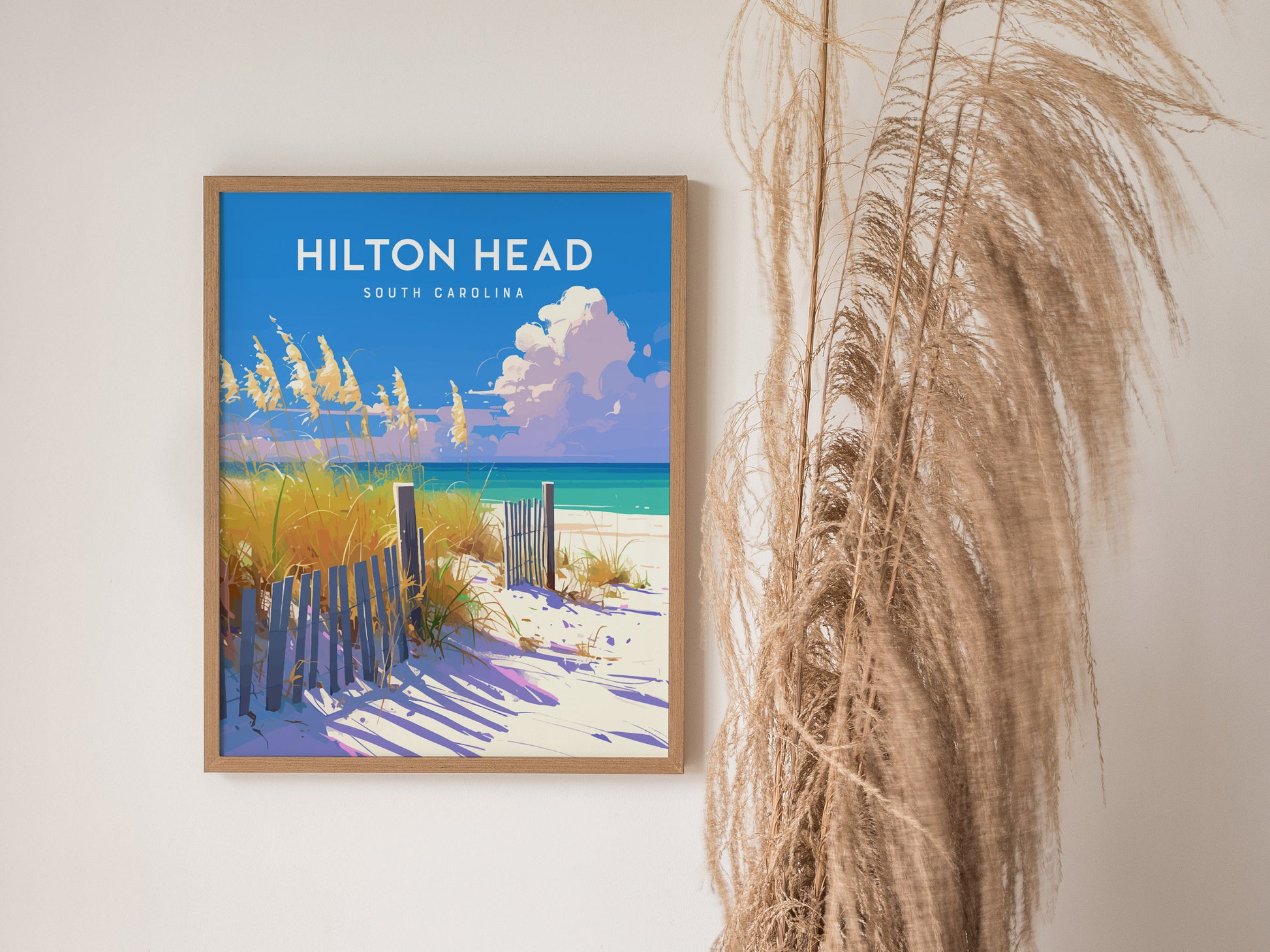 A framed poster of Hilton Head, South Carolina, depicts a sandy beach with a wooden fence, tall grasses, and a vibrant blue sky with fluffy clouds. The artwork is displayed on a white wall next to dried pampas grass.