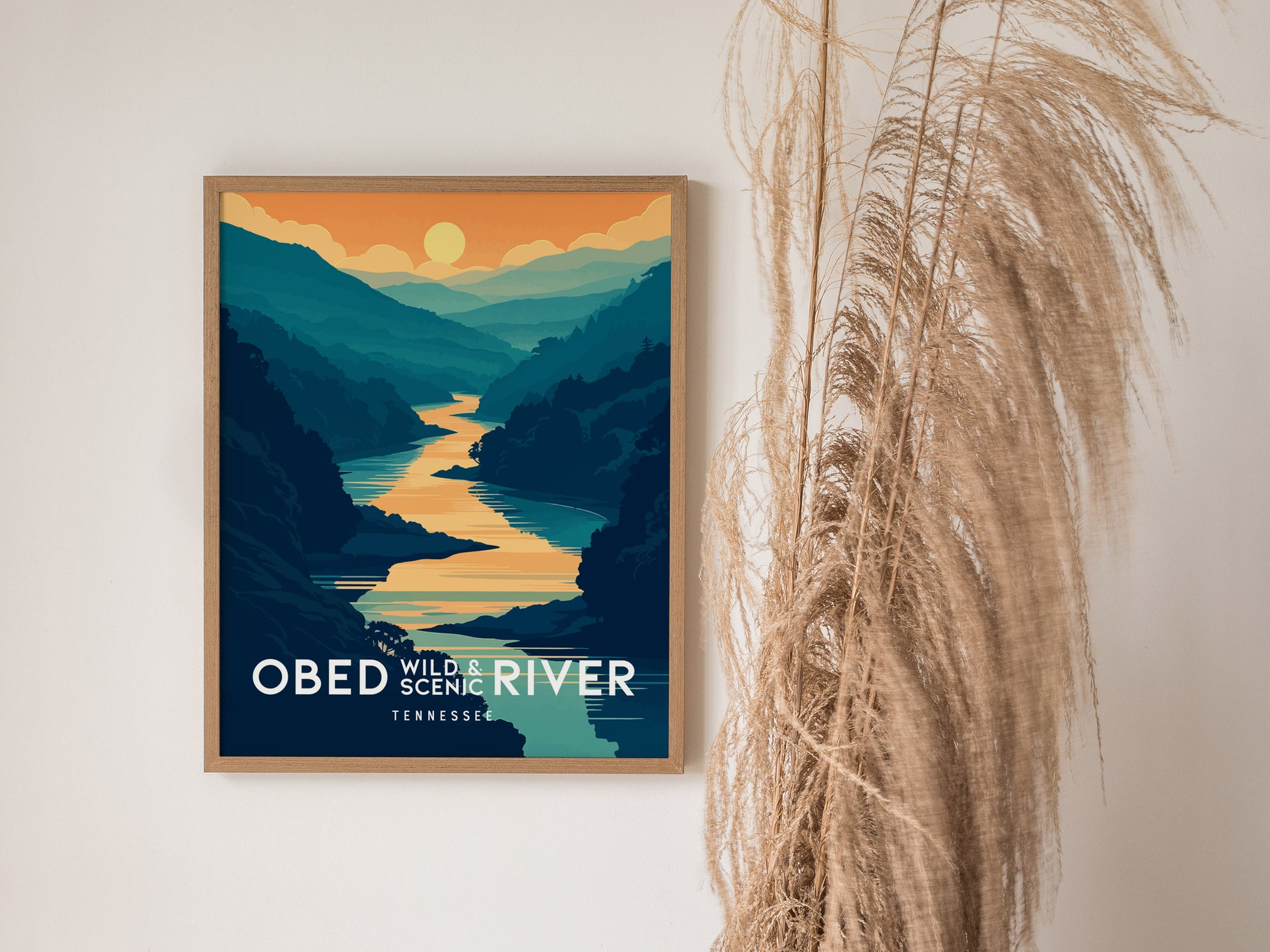 A framed poster depicting the Obed Wild and Scenic River with a vibrant sunset over layered mountains. It is placed on a wall beside tall, dried grasses.