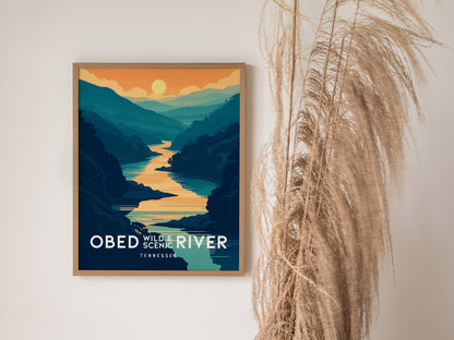 A framed poster depicting the Obed Wild and Scenic River with a vibrant sunset over layered mountains. It is placed on a wall beside tall, dried grasses.