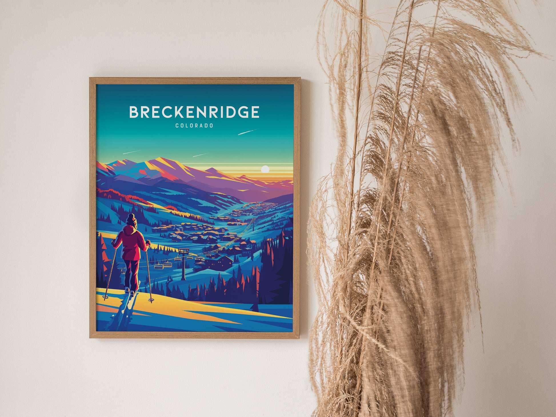 A framed poster of Breckenridge, Colorado, depicts a vibrant sunset over snow-covered mountains and a skier. Tall, dried grasses are positioned beside the artwork.