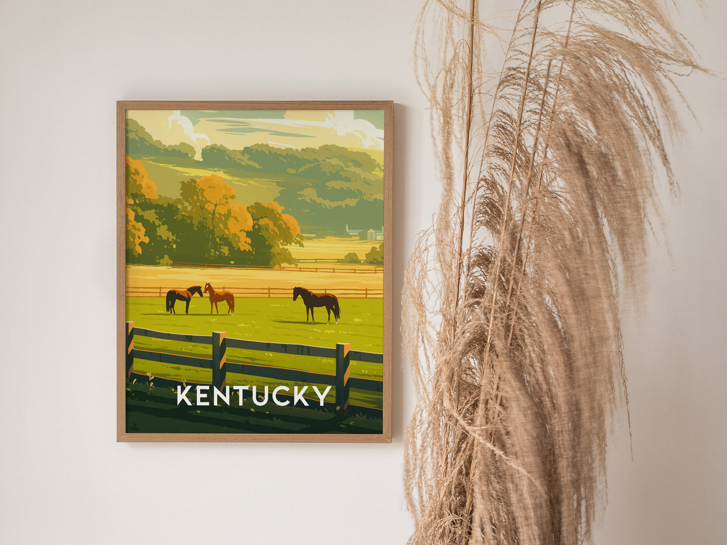 A framed poster depicting horses grazing in a lush, green Kentucky pasture with a wooden fence and vibrant autumn trees in the background, next to dried pampas grass.