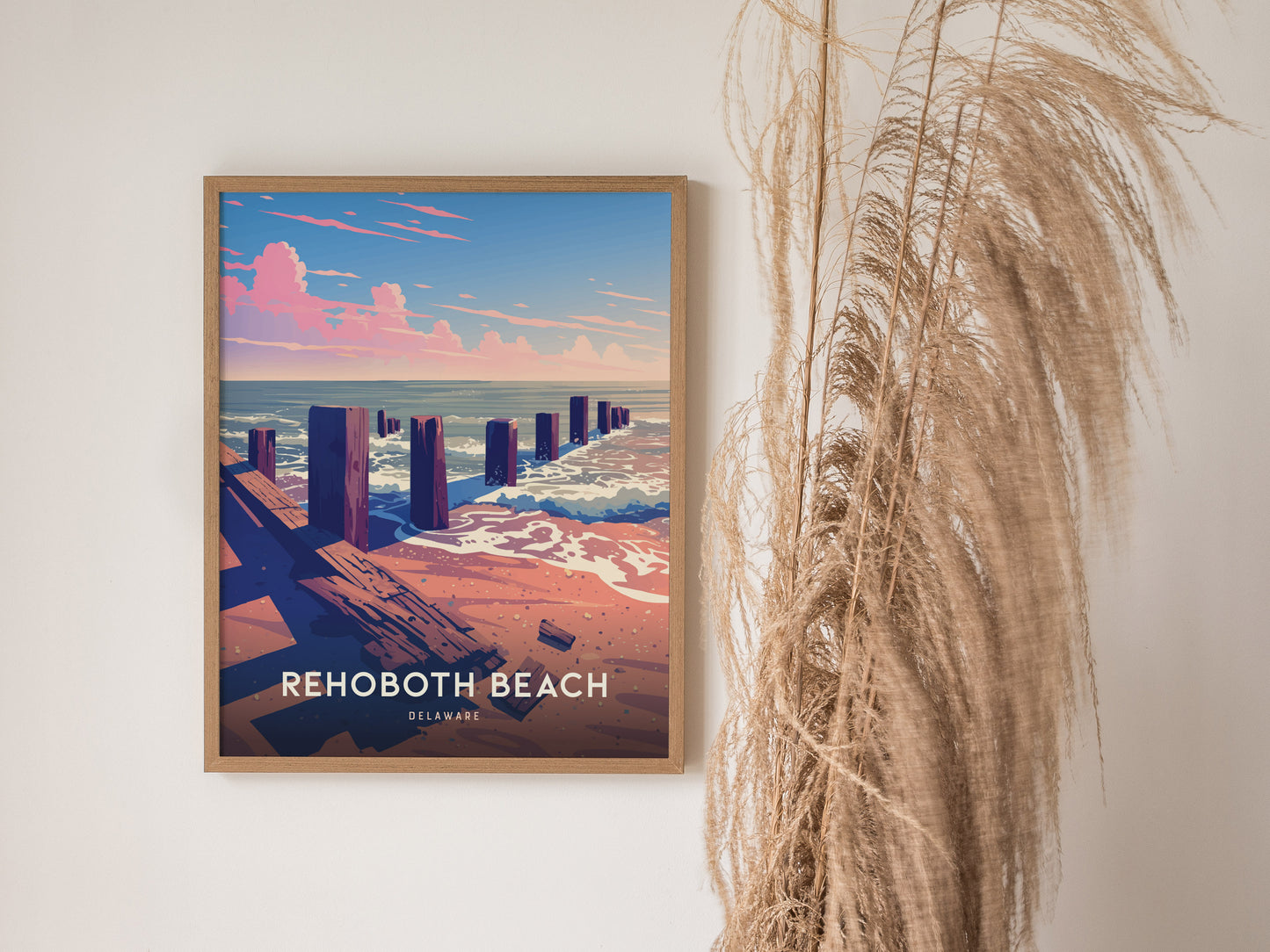 A framed poster of Rehoboth Beach featuring a stylized boardwalk and ocean scene with vibrant pink and blue skies, displayed next to tall, dried plants.