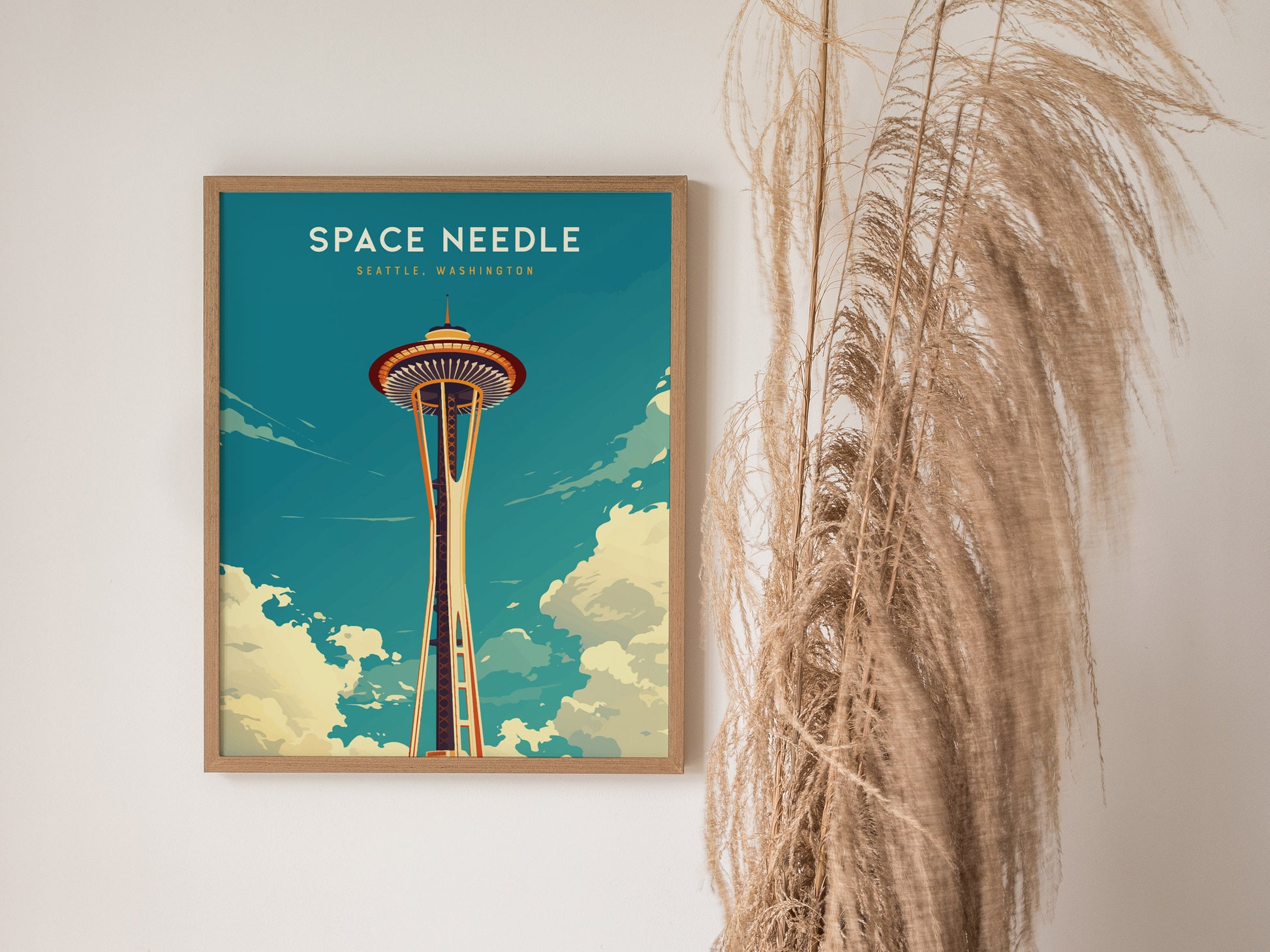 Framed Space Needle poster in vintage style hangs on a wall, accompanied by tall dried pampas grass. The artwork showcases Seattles iconic tower against a blue and cloudy sky.