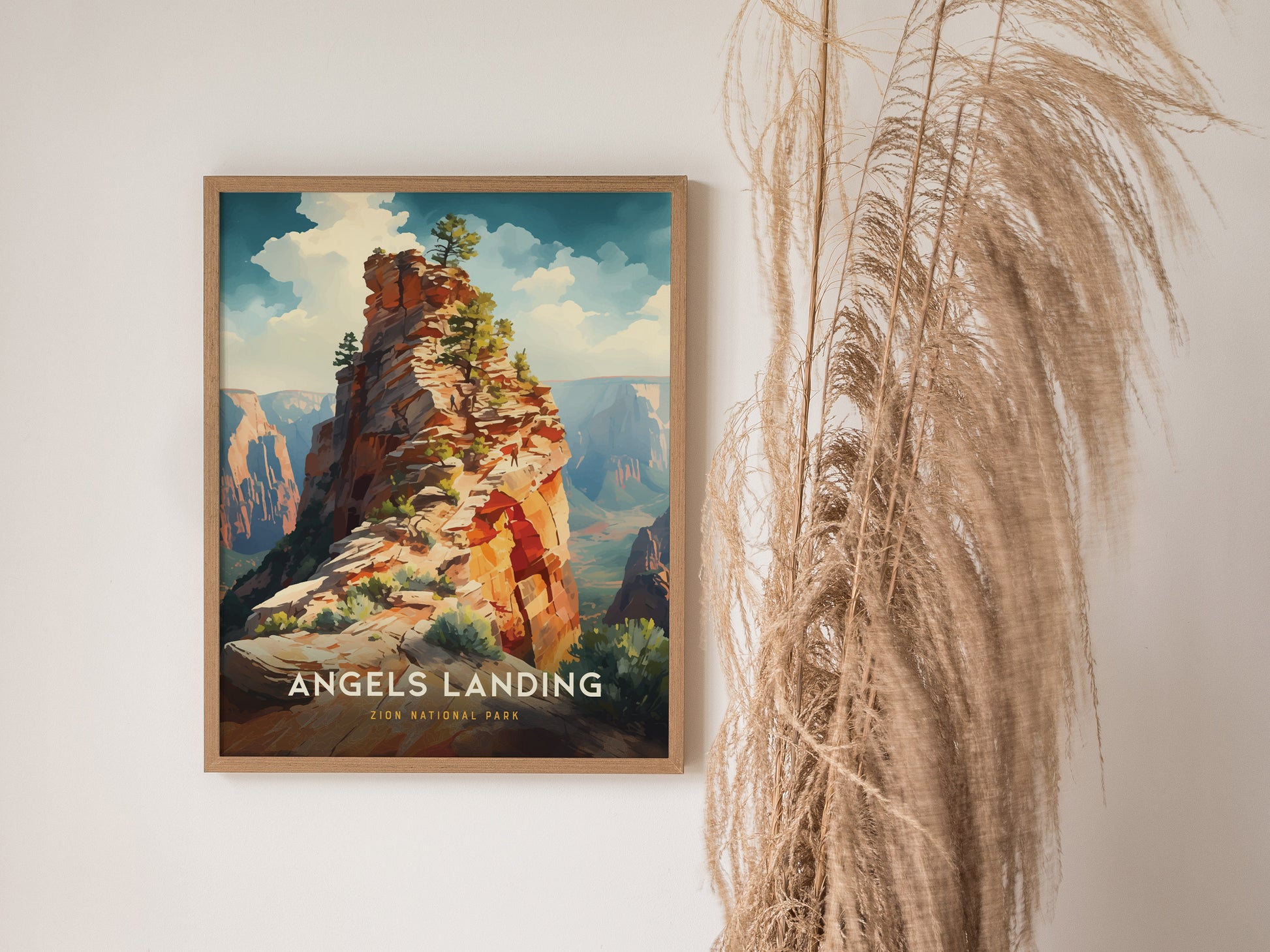 A framed poster of Angels Landing in Zion National Park hangs on a white wall beside tall, wispy pampas grass. The artwork features a vibrant depiction of the sandstone ridge.