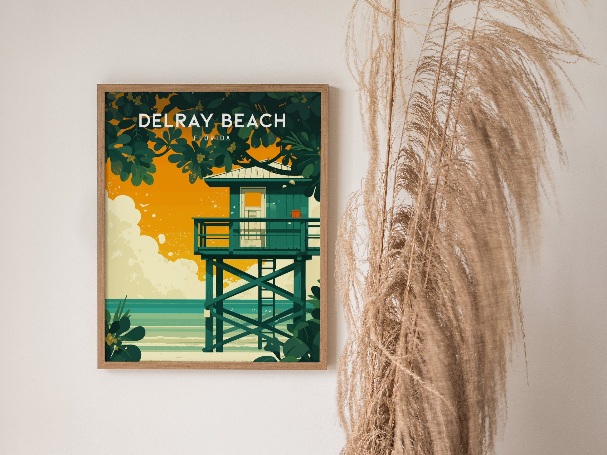 A framed Delray Beach poster features a lifeguard stand against a vibrant orange sky, surrounded by lush greenery. Tall dried grasses are placed beside it for decoration.
