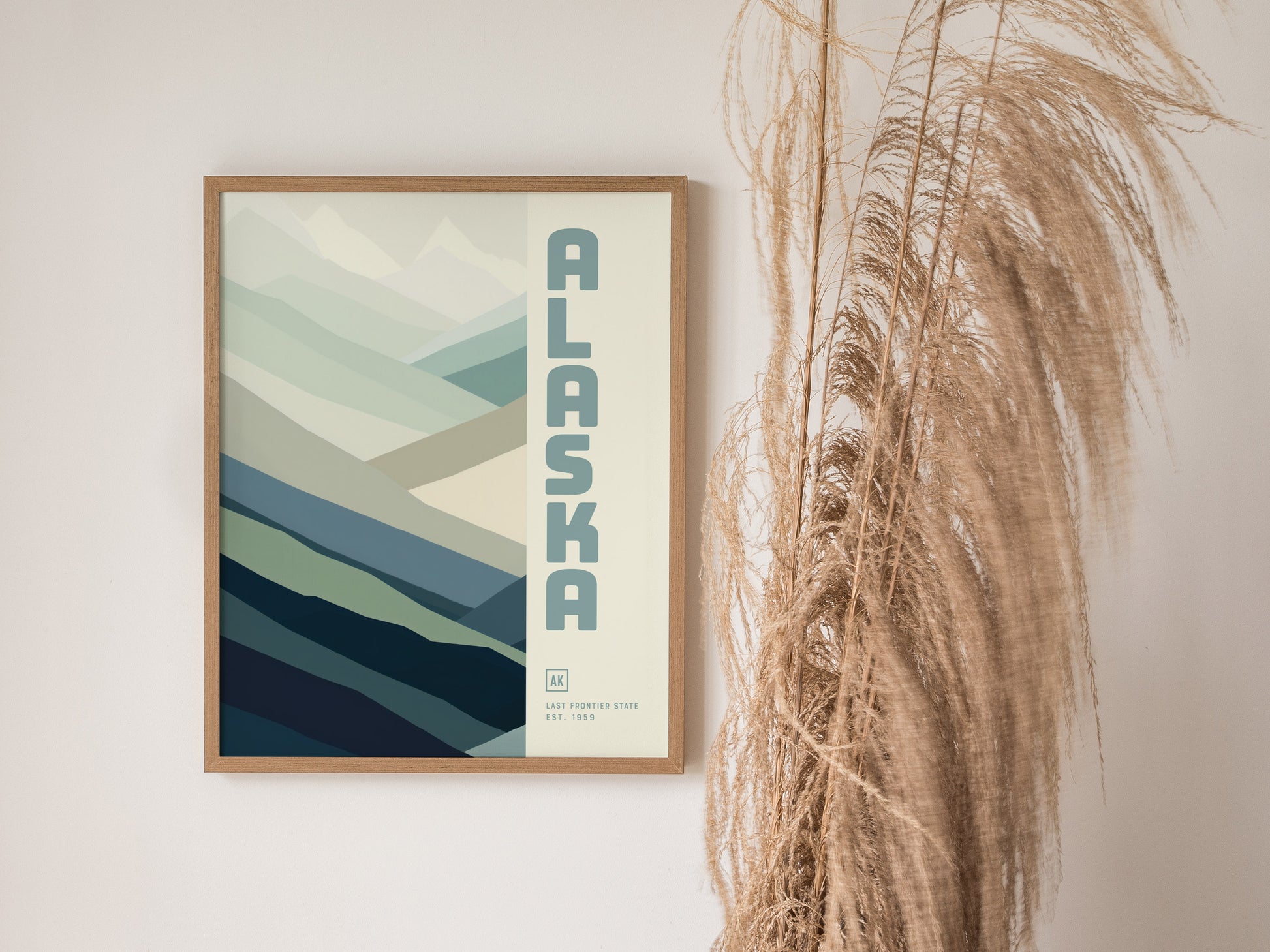 A framed Alaska poster with a minimalist mountain design and blue-green tones is displayed on a wall next to tall, wispy dried grasses.
