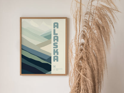 A framed Alaska poster with a minimalist mountain design and blue-green tones is displayed on a wall next to tall, wispy dried grasses.