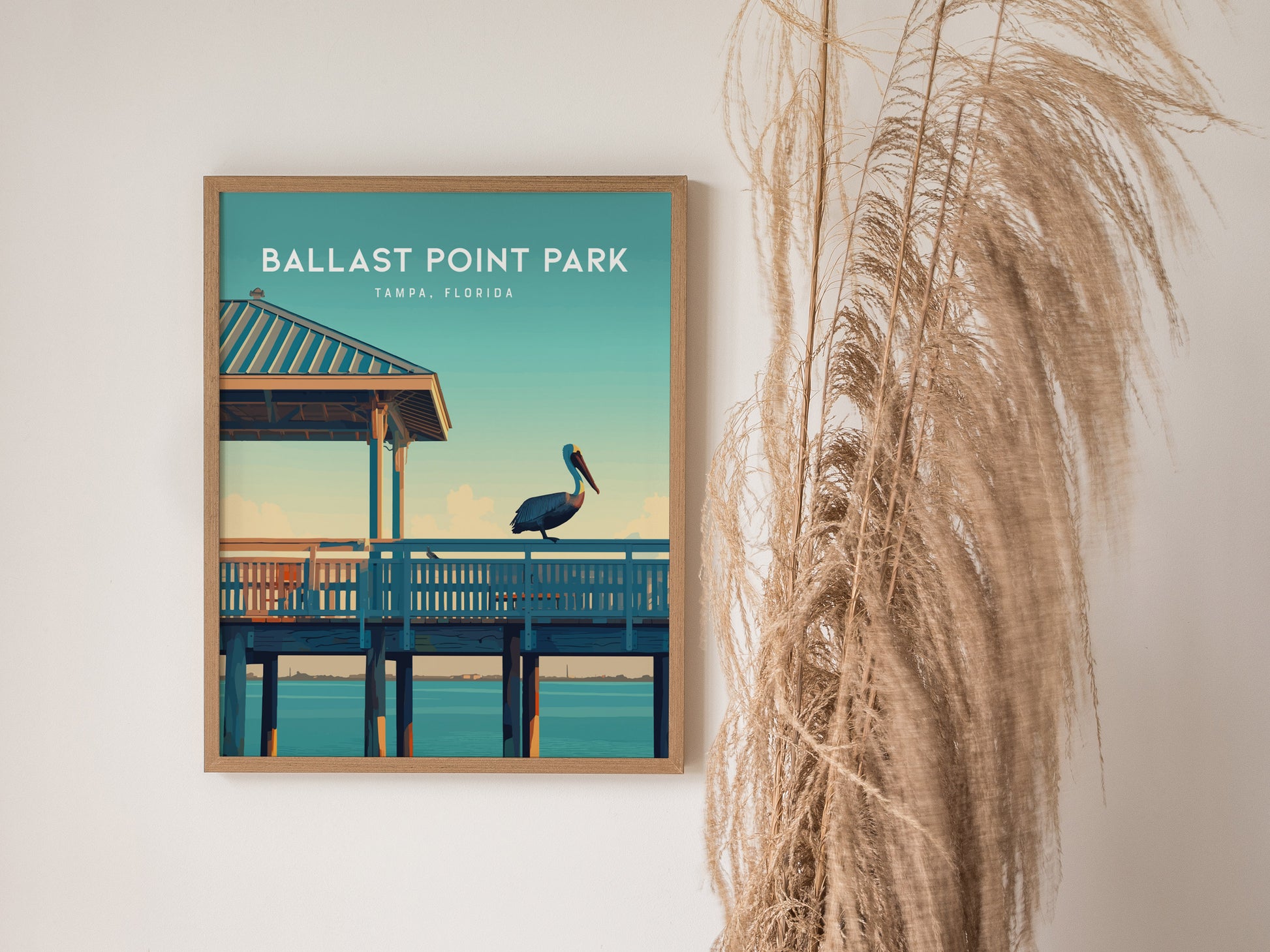 A framed poster of Ballast Point Park, Tampa, Florida, featuring a pelican on a pier, is displayed beside tall dried grasses against a white wall.