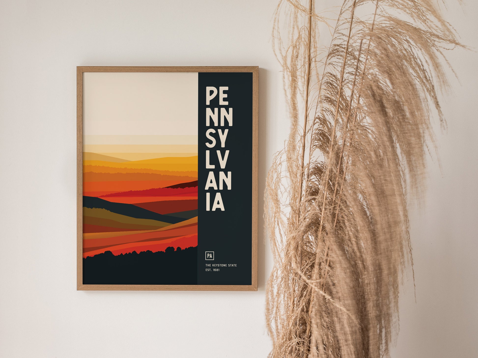 A framed poster featuring an abstract landscape design in warm tones with the word PENNSYLVANIA vertically aligned on a black strip. Dried pampas grass decorates the right side.