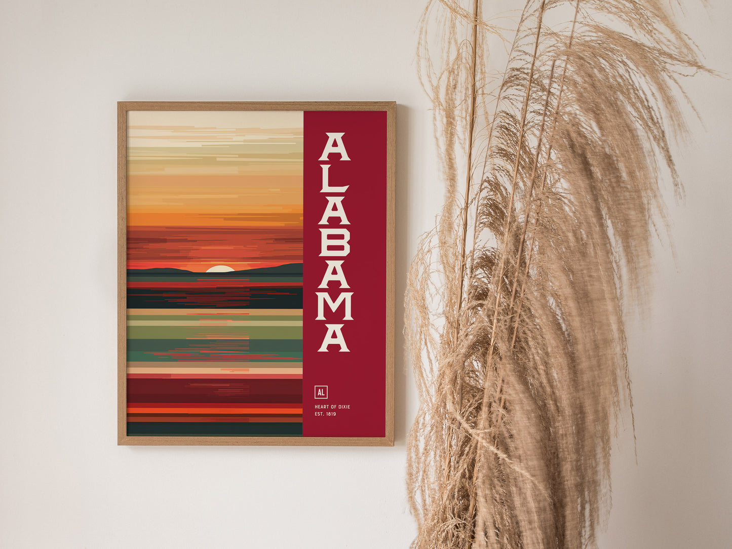 A framed abstract poster with Alabama in bold white letters on a red background, depicting a colorful sunset landscape. Tall dried grasses are visible beside the artwork.