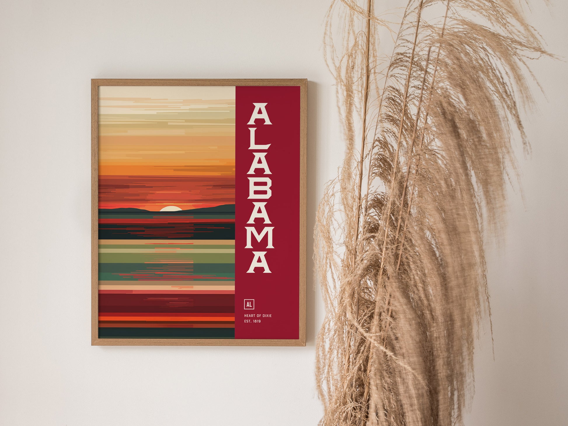 A framed abstract poster with Alabama in bold white letters on a red background, depicting a colorful sunset landscape. Tall dried grasses are visible beside the artwork.