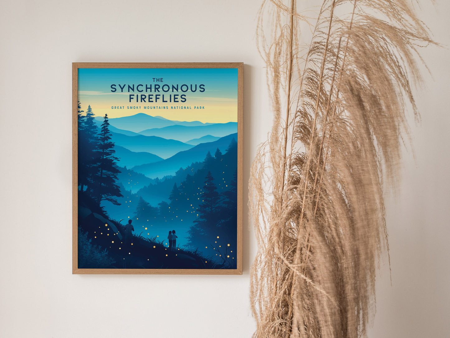 A framed poster titled The Synchronous Fireflies depicts a scenic view of the Great Smoky Mountains with glowing fireflies and silhouettes of people. Its mounted on a wall beside tall, dried grasses.