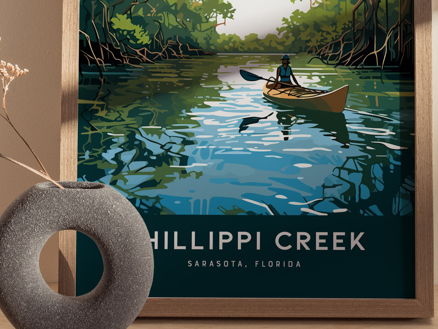 A framed poster depicts a kayaker paddling on a serene creek surrounded by lush mangroves under a clear sky. The text reads Phillippi Creek, Sarasota, Florida.