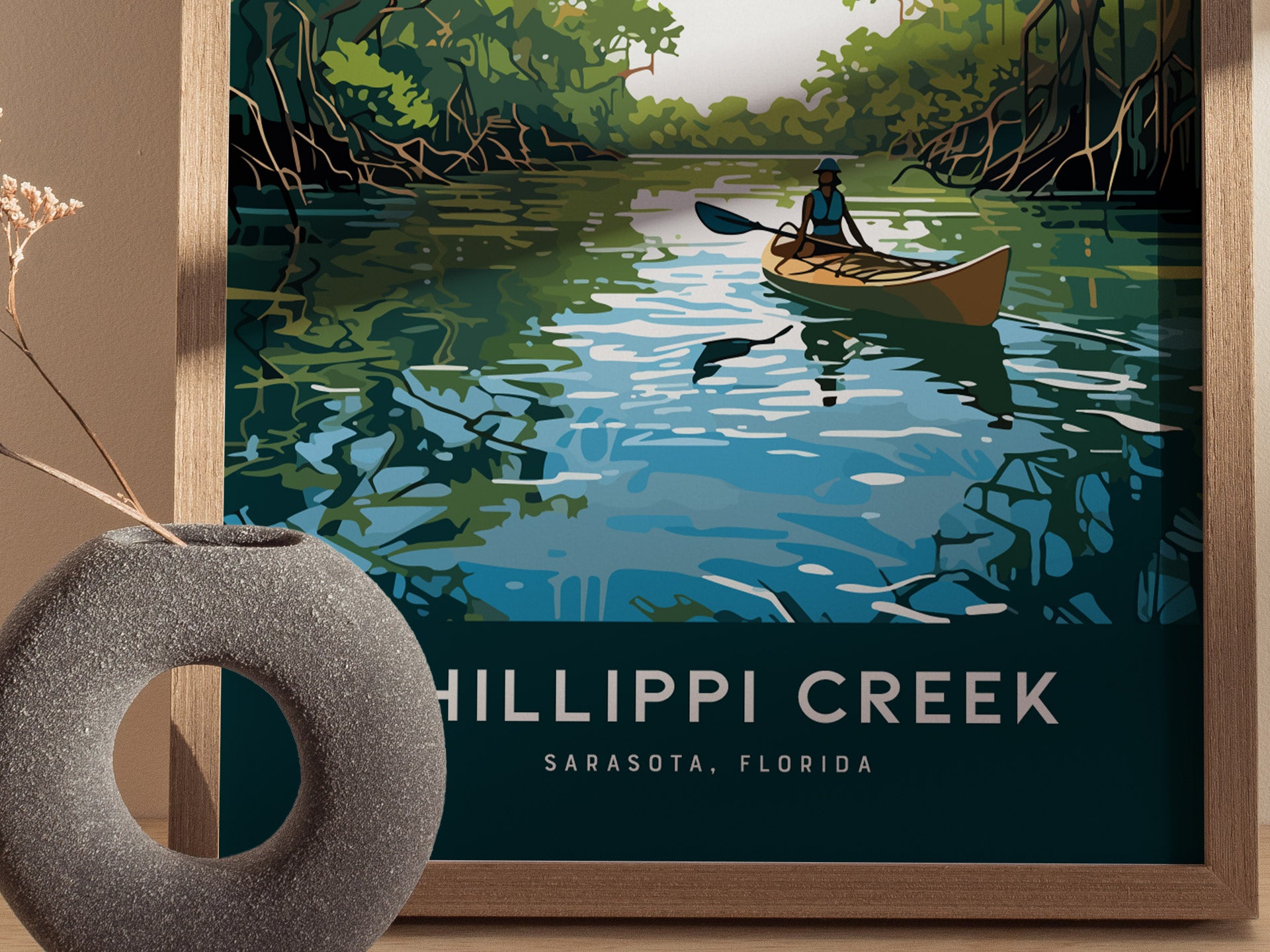 A framed poster depicts a kayaker paddling on a serene creek surrounded by lush mangroves under a clear sky. The text reads Phillippi Creek, Sarasota, Florida.