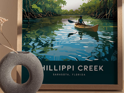 A framed poster depicts a kayaker paddling on a serene creek surrounded by lush mangroves under a clear sky. The text reads Phillippi Creek, Sarasota, Florida.