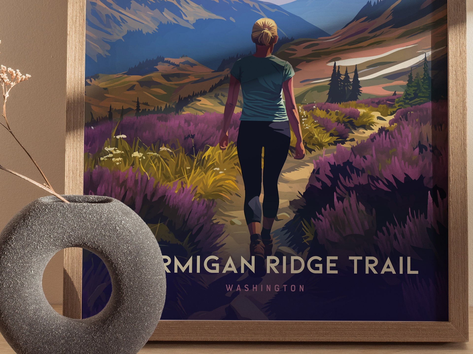 A framed poster depicts a woman hiking along a trail surrounded by vibrant wildflowers and mountains. A round, textured vase with dried flowers is placed nearby.