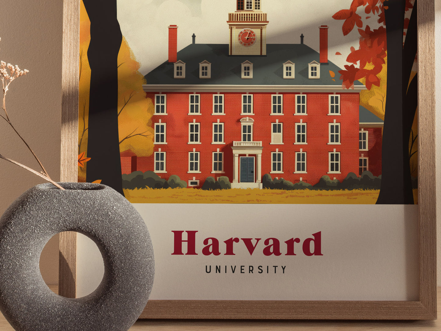 Framed poster of a red-brick building with a clock tower, surrounded by autumn trees, labeled Harvard University. A gray circular vase with dried flowers is in the foreground.