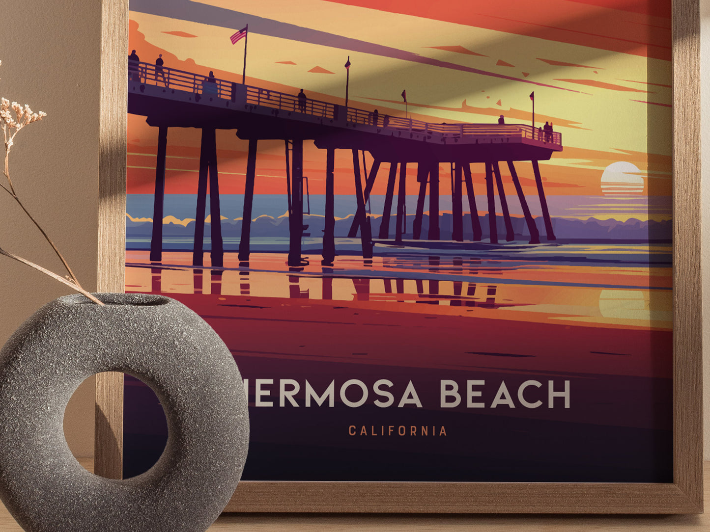 A framed poster of Hermosa Beach Pier at sunset features vibrant hues of orange, pink, and purple, with silhouetted figures on the pier. A round, textured vase sits beside it.