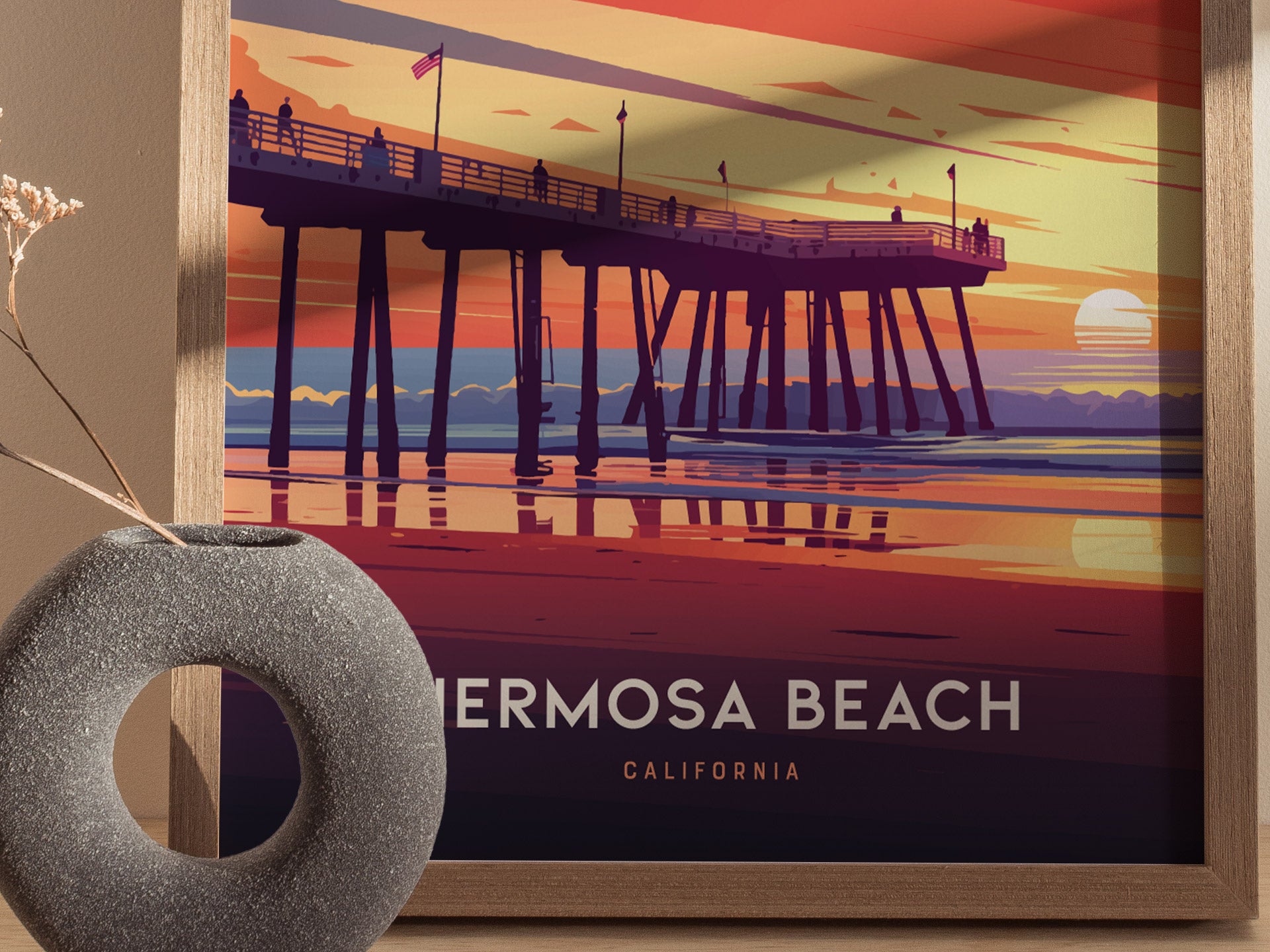 A framed poster of Hermosa Beach Pier at sunset features vibrant hues of orange, pink, and purple, with silhouetted figures on the pier. A round, textured vase sits beside it.