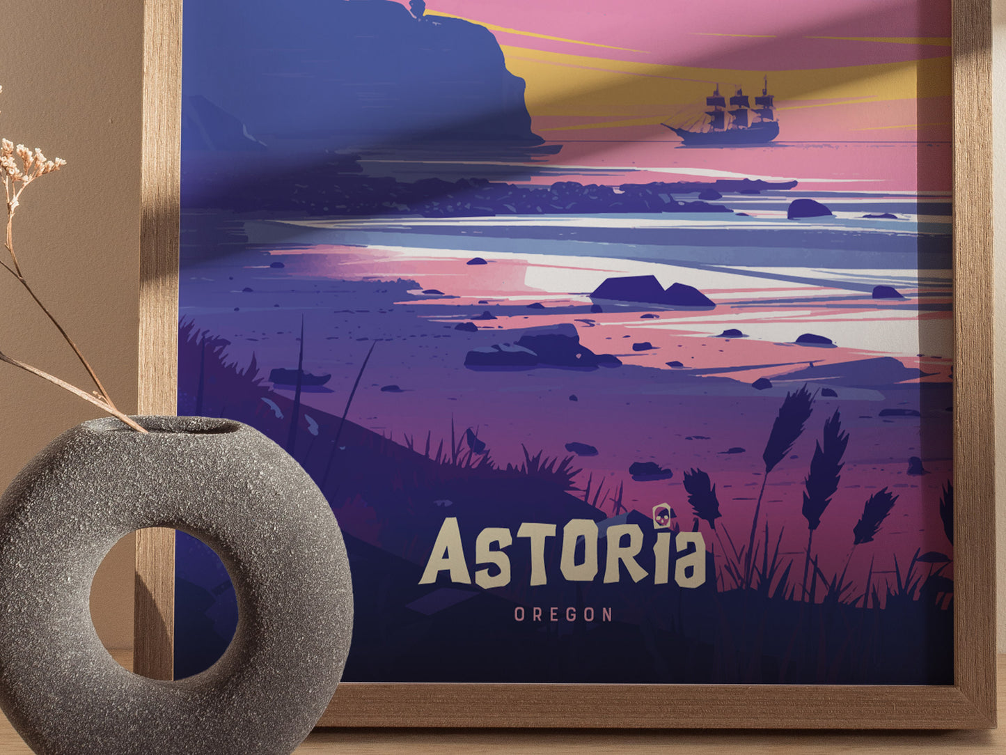 A framed poster shows a stylized sunset over a rocky beach with a pirate ship in the distance, featuring the text Astoria Oregon. A round stone vase sits nearby.