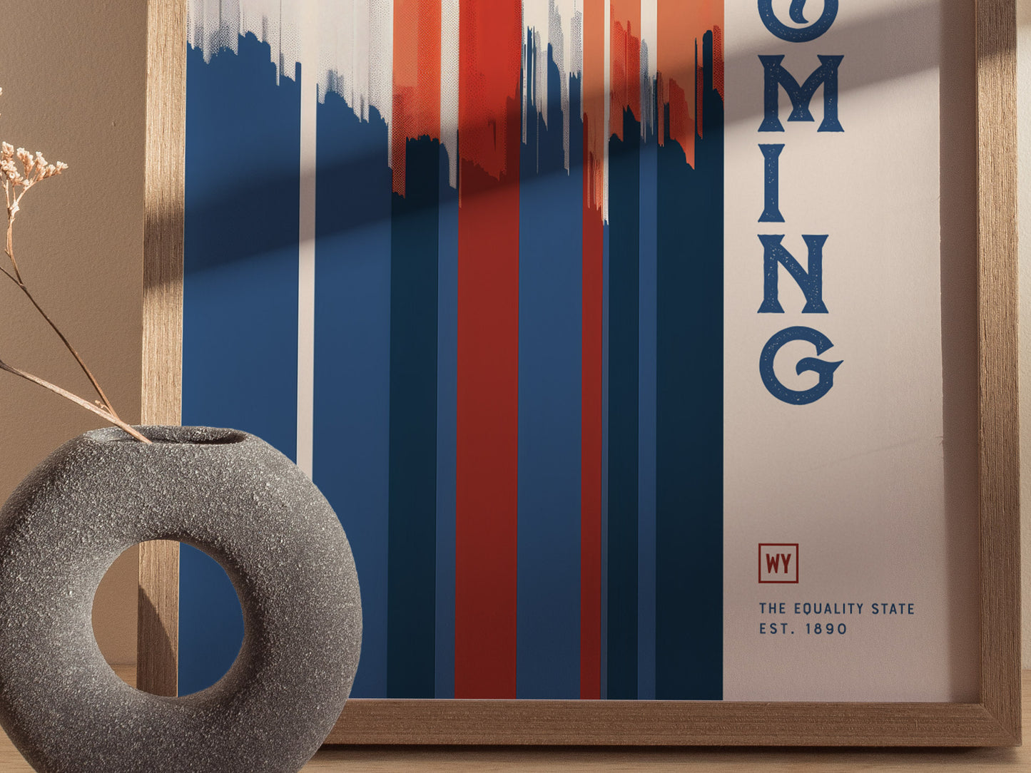 A framed Wyoming poster features red, white, and blue vertical geometric shapes with the text The Equality State Est. 1890. A textured circular vase with dried flowers is beside it.
