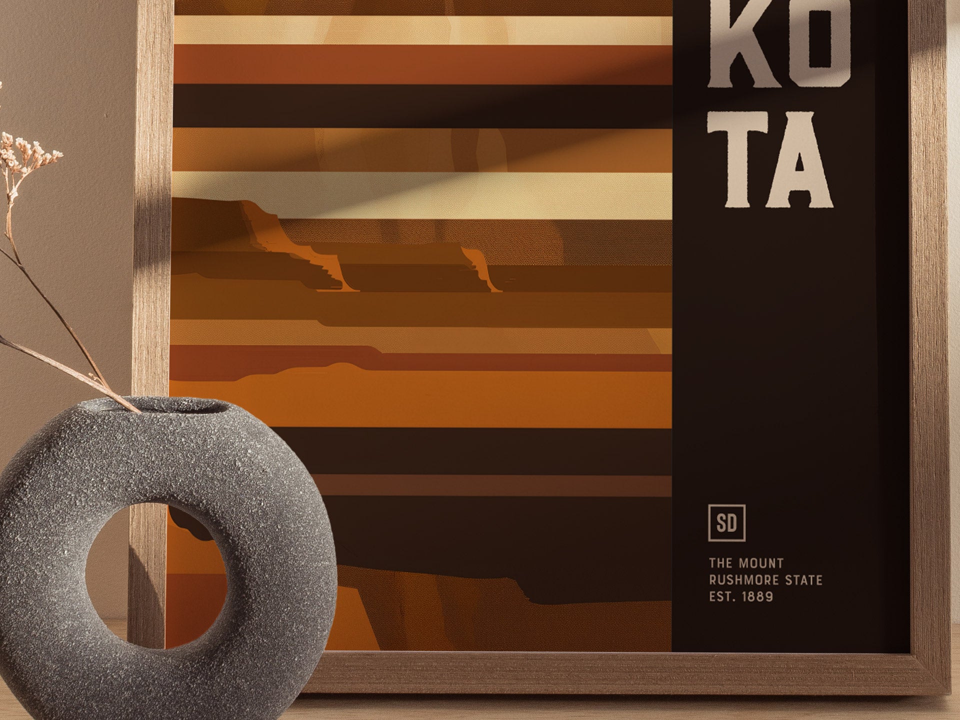 A minimalist geometric poster features earthy tones and abstract mountain shapes, partially framed in wood, with the words KOTA visible. A textured, circular vase with dried flowers sits nearby.