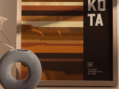 A minimalist geometric poster features earthy tones and abstract mountain shapes, partially framed in wood, with the words KOTA visible. A textured, circular vase with dried flowers sits nearby.