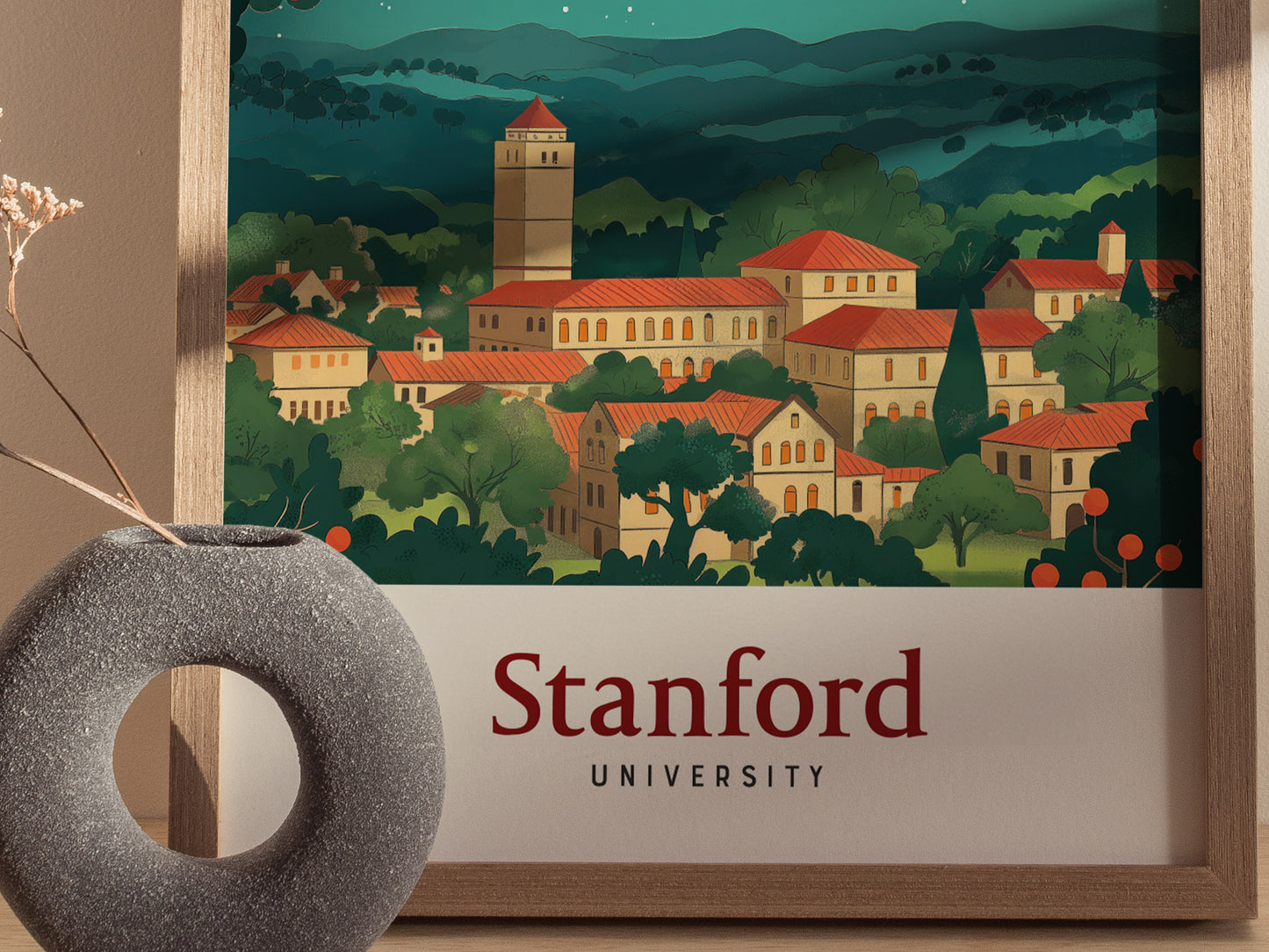 A vintage-style poster of Stanford University features red-roofed buildings amidst lush greenery, framed in wood. A modern gray circular vase with dried flowers is in the foreground.