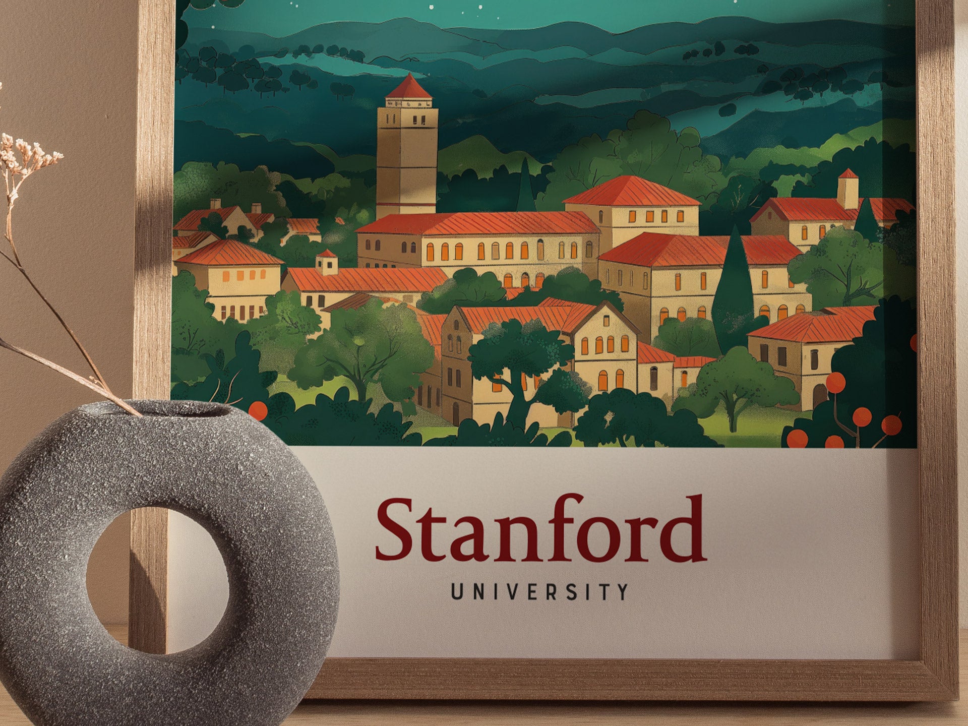 A vintage-style poster of Stanford University features red-roofed buildings amidst lush greenery, framed in wood. A modern gray circular vase with dried flowers is in the foreground.