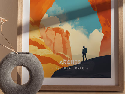 A minimalist poster of Arches National Park features a lone hiker beneath towering red rock formations and a vibrant sky. A round stone vase with dried flowers sits nearby.