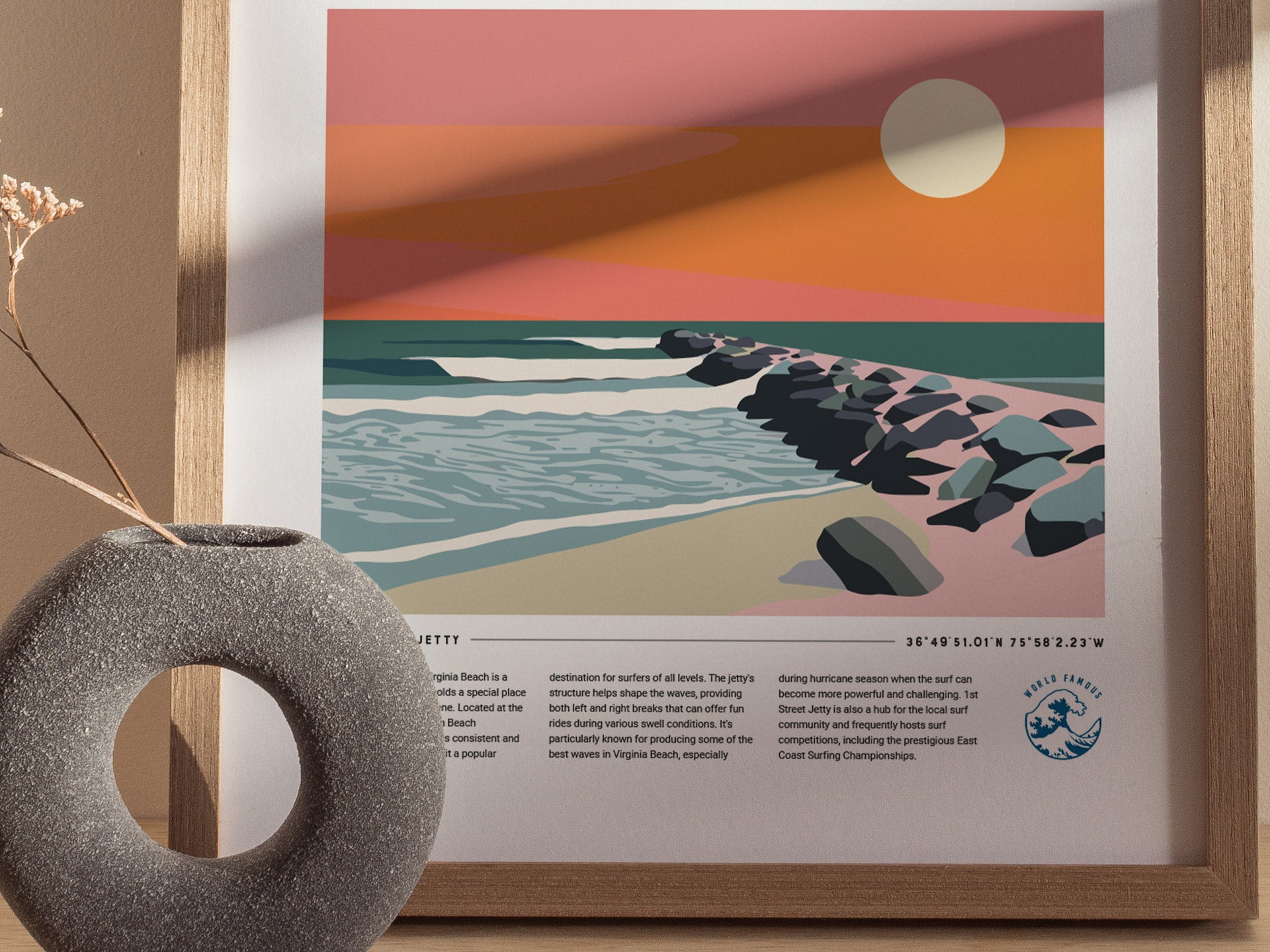 A framed poster of a colorful beach scene featuring a rocky jetty, waves, and a setting sun. A textured circular vase with a dried plant is in the foreground.