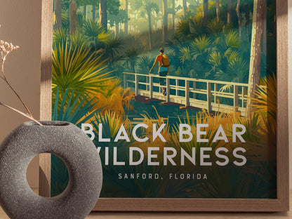A framed poster of a person on a wooden bridge in a lush forest, with Black Bear Wilderness, Sanford, Florida text, sits beside a textured round vase.