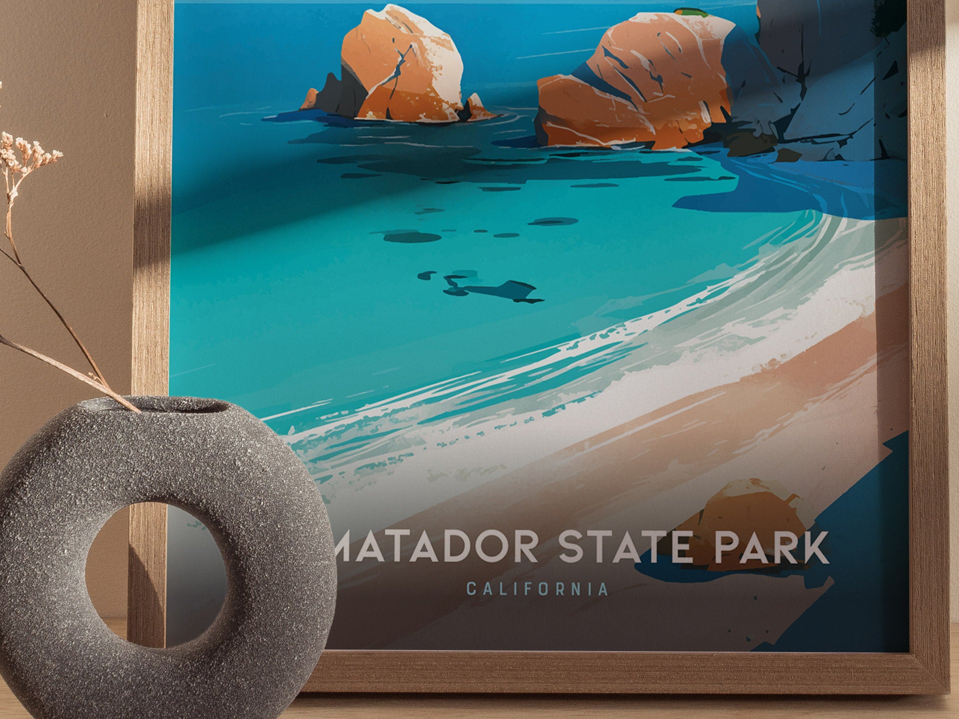 A framed poster of El Matador State Park showcases vibrant turquoise waters, golden sand, and rugged cliffs. A round stone vase with dried flowers is also visible in the foreground.