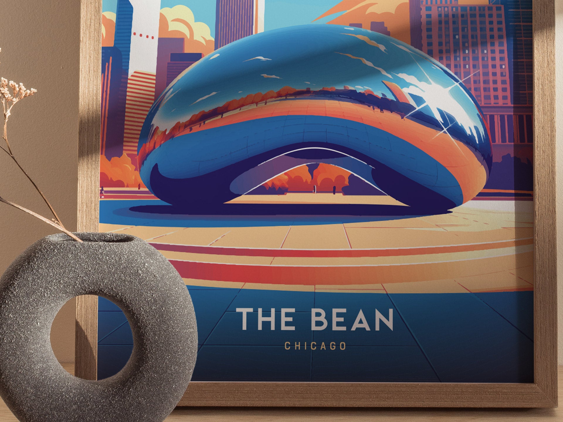 A framed poster of The Bean in Chicago, featuring colorful reflections and surrounding skyscrapers, is displayed beside a round, textured vase with dried branches.