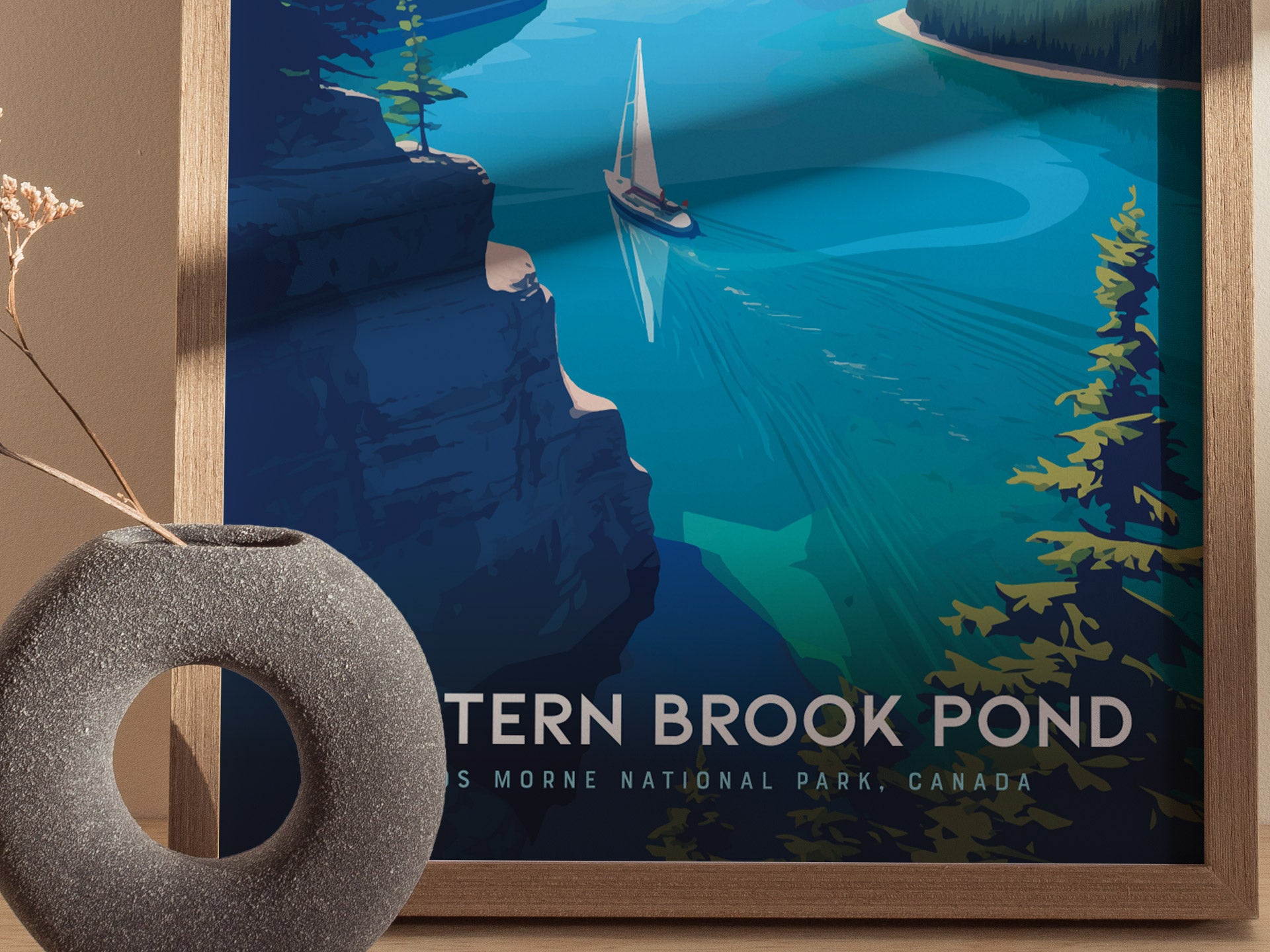 A framed poster of Western Brook Pond depicts a sailboat gliding through vibrant blue waters surrounded by towering cliffs and lush greenery. A textured, round vase is in the foreground.