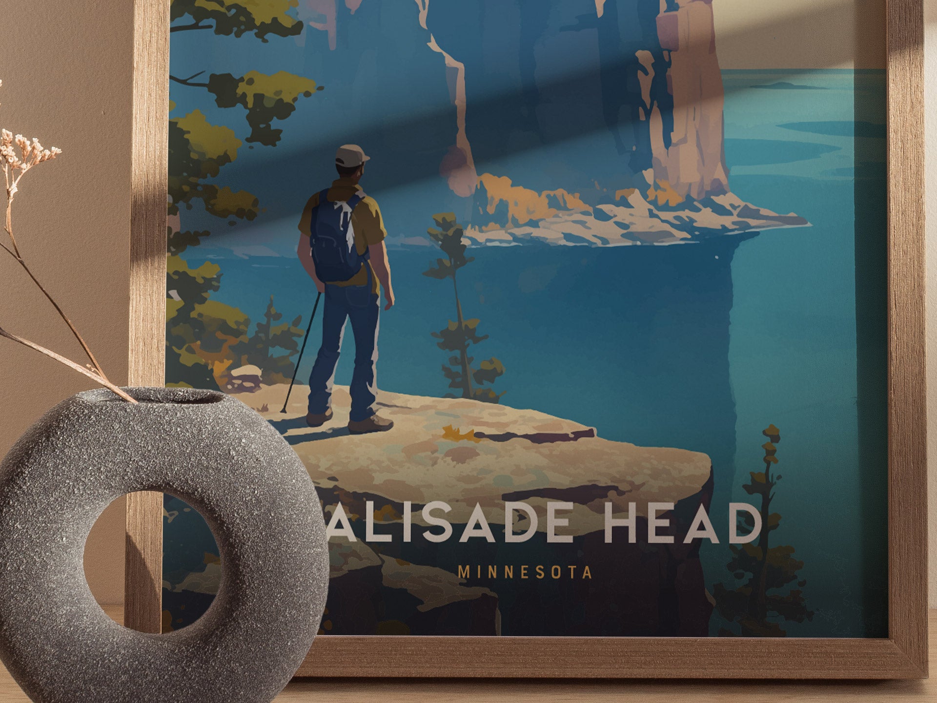 A framed poster depicts a hiker with a backpack standing on a cliff overlooking a lake, surrounded by trees. A gray vase with dried flowers is in the foreground.
